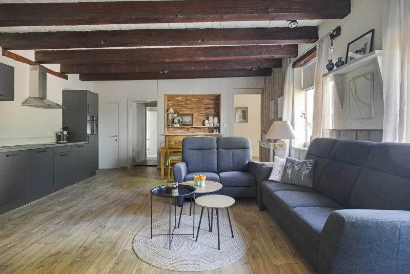 De moderne woonkamer van het 4-6 persoon vakantiehuis te huur bij Paal Zeeland met een open keuken, houten vloeren, een bank, een fauteuil en een salontafel, ramen met witte gordijnen.