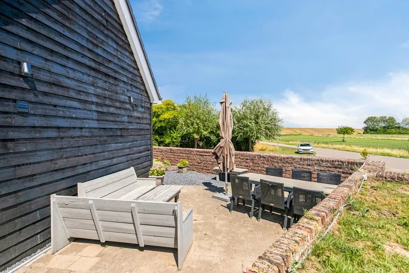Buitenruimte met een houten huis, tuinmeubilair, een parasol en een omheind terras met uitzicht op velden en bomen van het 2 persoon vakantiehuis te huur bij Paal.