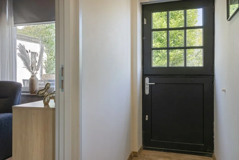 Ingang van het vakantiehuis te huur bij Paal nabij Hulst met zwarte deur met ruit, witte muur en gedeeltelijk zicht op woonkamer met raam, kast en decoratieve voorwerpen.