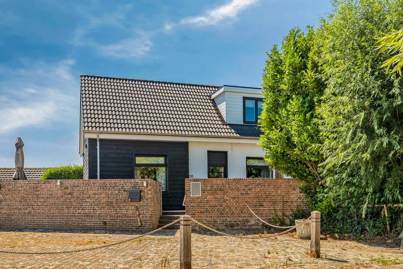 Moderne witte en zwarte vakantiehuis te huur met stenen muur en grote groene boom op de rechterkant, onder een blauwe lucht bij Paal, Zeeuws-Vlaanderen 