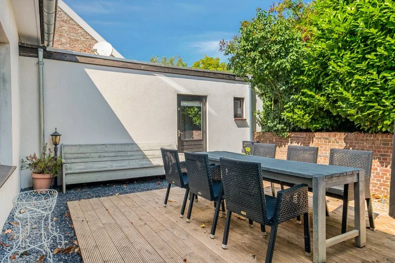 Buiten eethoek met zwart tafel en zes zwart wicker stoelen, houten terras, groene struik en witte muur, terrasverlichting, zonnige dag.