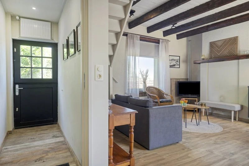 Interieur van het 2 persoon vakantiehuisje bij Paal en de woonkamer met een bank, een fauteuil, televisiescherm, ramen met gordijnen en houten plafondbalken.