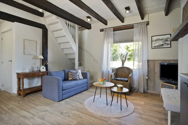 Woonkamer van het 2 persoon vakantiehuisje in de buurt van Kloosterzande met houten vloer, blauwe bank, rotanstoel, tv, witte gordijnen voor raam, open trap, en plafond met houten balken.