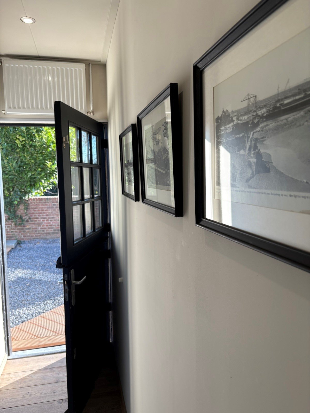 Hal met openstaande deur, zwart-wit foto's aan de muur en zicht op een tuin met grind en houten vlonder.