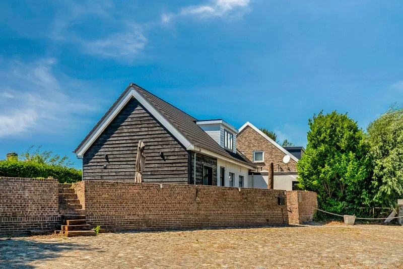 Het vakantiehuis Den Burtman bij Paal te huur voor 2 - 8 personen met een bruin bakstenen muur, groene bomen, en een blauwe hemel met enkele wolken.