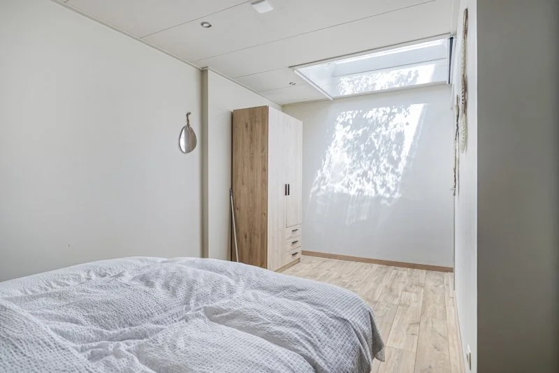 Een eenvoudige slaapkamer met een wit bed en een houten kast, met veel natuurlijk licht dat door een dakraam naar binnen valt.