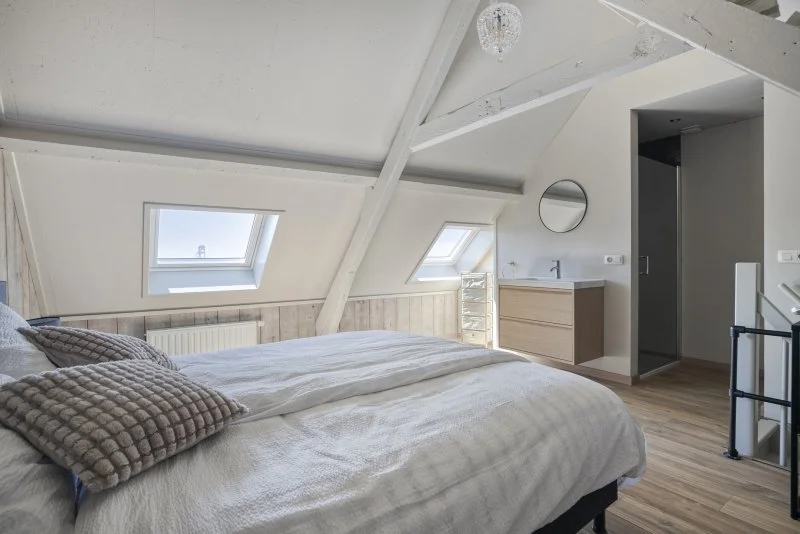 De slaapkamer met een grote bed, kussens, twee dakramen, een spiegel, een wastafel met kastje, en een deur naar de badkamer van het 2 persoon vakantiehuis te huur bij de Westerschelde gemeente Hulst.