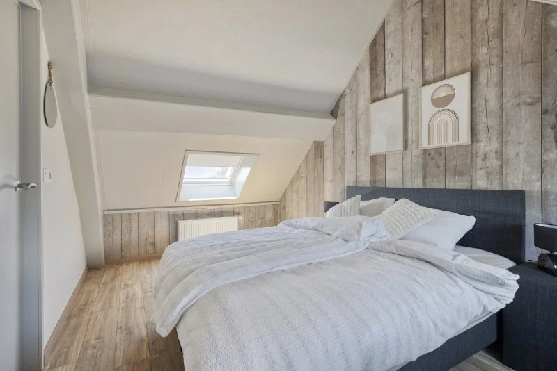 Een slaapkamer met een bed, witte beddengoed en kussens, houten muurdecoraties, een raam in het plafond, en een houten vloer.
