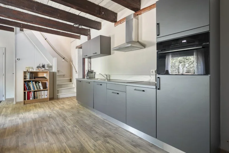 Moderne keuken met grijze kasten, houten vloer en een open trap met boekenplank in de woonkamer.