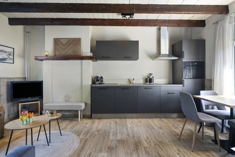 Modern keuken met donkere kasten, houten vloer en zitgedeelte, glazen deur met gordijn aan de rechterkant.