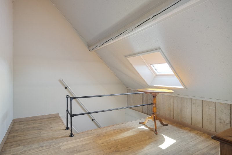 Een klein hoekje van een zolderkamer met een houten tafel en een raampje in het schuine plafond dat daglicht binnenlaat.