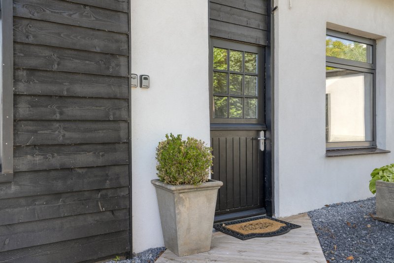 De afbeelding toont de ingang van het vakantiehuis met een zwarte deur, een lichtgrijze muur, en een tuin met planten in potten van het 4-6 persoon vakantiehuis te huur bij Paal.