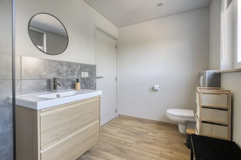 Een minimalistische badkamer met wastafel, spiegel, toilet en raam, lichte muren en houten vloer.