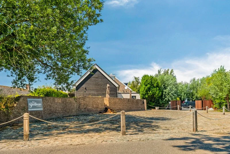 Parkeerplaatsen met het vakantiehuis Den Burtman op de achtergrond omgeven door bomen en een openluchtterrein met een hek van houten palen en touwen in Kruispolderhaven, bij Paal, hulst en Kloosterzande.
