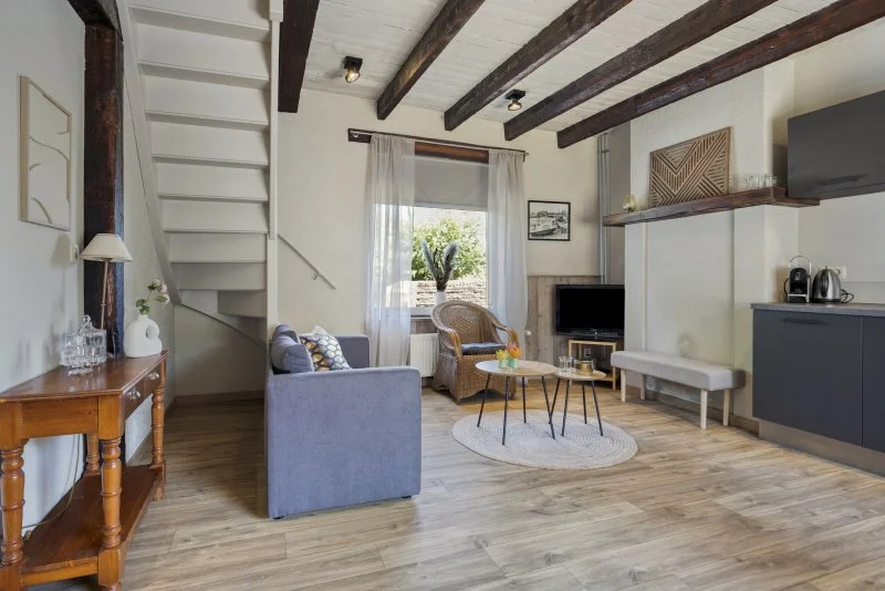 Woonkamer van het 2 persoon vakantiehuisje bij Paal met houten vloeren, donkere houten plafondbalken, een blauw bankje, een rieten stoel, een tv en een raam met witte gordijnen die uitzicht bieden op de tuin.