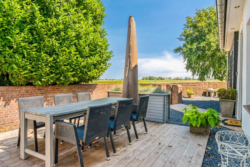 Een buiten eethoek met zes stoelen, een grote parasol, tuinplanten, een houten terras, een muur van bakstenen en een open landschap met bomen en een blauwe lucht.