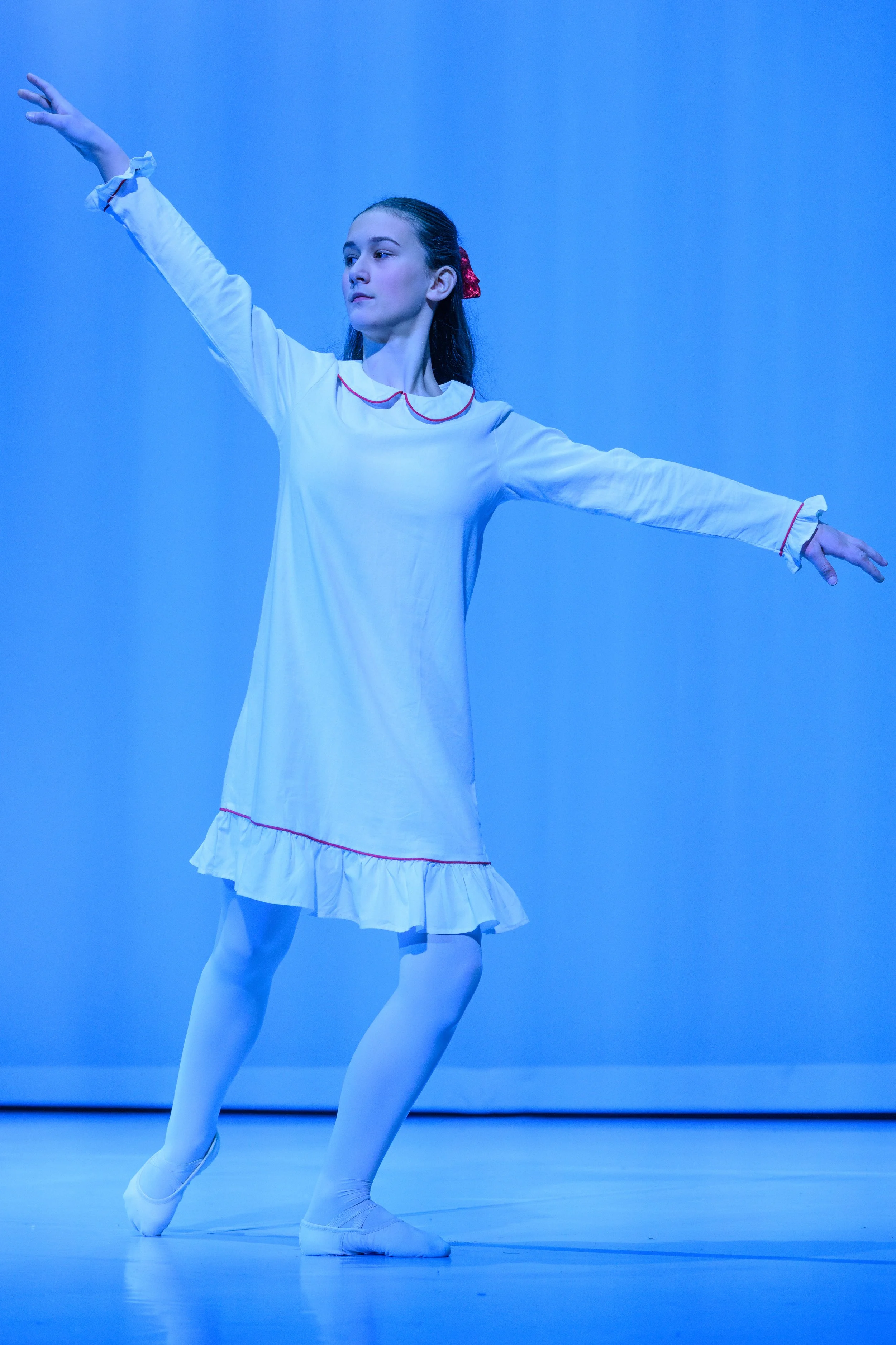 A young female ballet dancer in a white dress with red trim, performing a dance move on stage with a blue background.