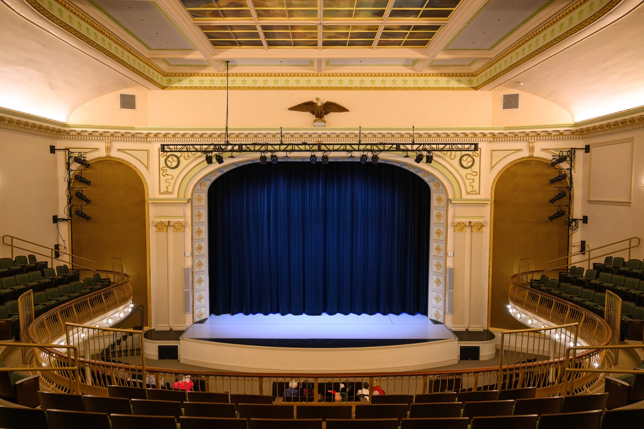 An ornate theater with a stage covered by a blue curtain, golden railings, and seating surrounding the stage in a semi-circle, with warm lighting.