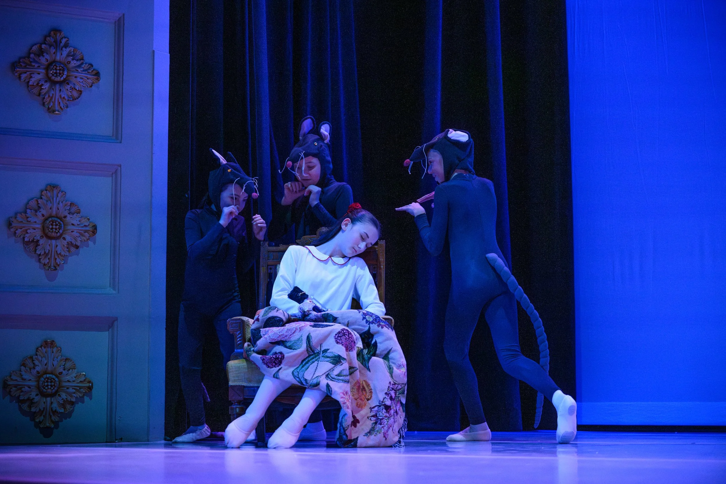 A stage scene from the ballet 'The Nutcracker' with a young woman in a white top and floral skirt sitting with her eyes closed, surrounded by four performers dressed as mice in black costumes with mouse ears and tails, with a dark curtain backdrop.