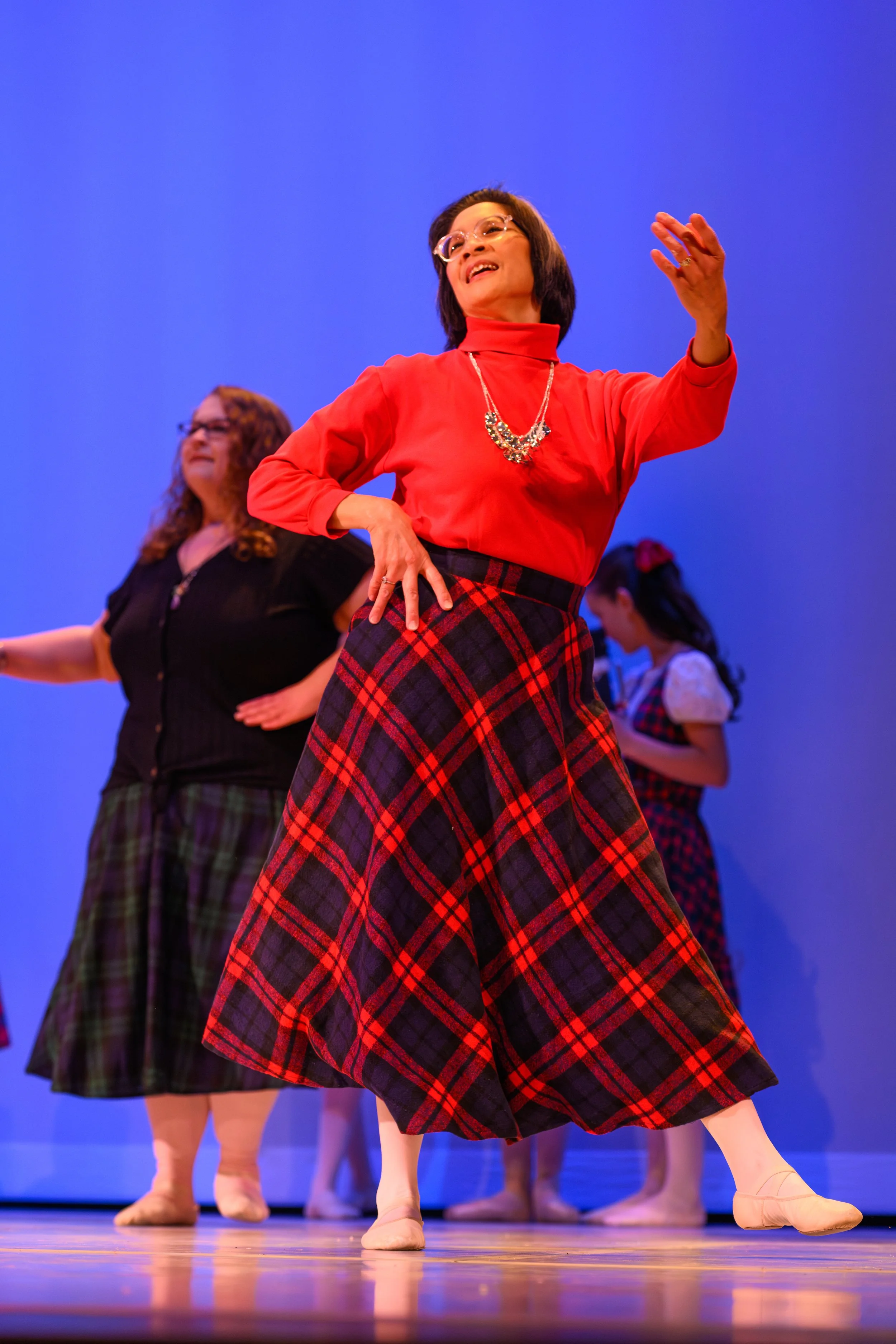 A woman wearing glasses, a red turtleneck, and a red and black plaid skirt, dancing on stage with other women. The stage background is blue.