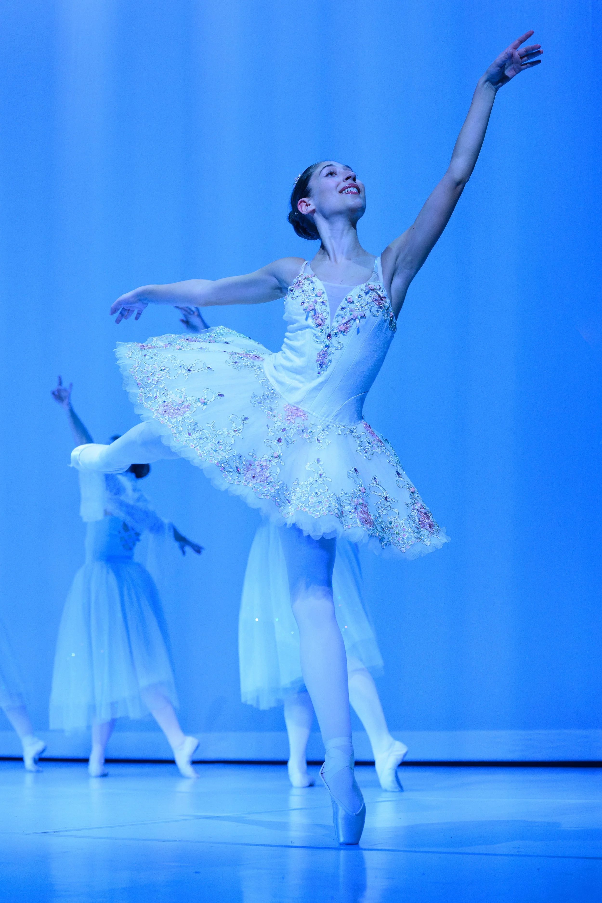 A ballerina in a white tutu dancing on stage with arms raised, surrounded by other dancers in white tutus, against a blue background.