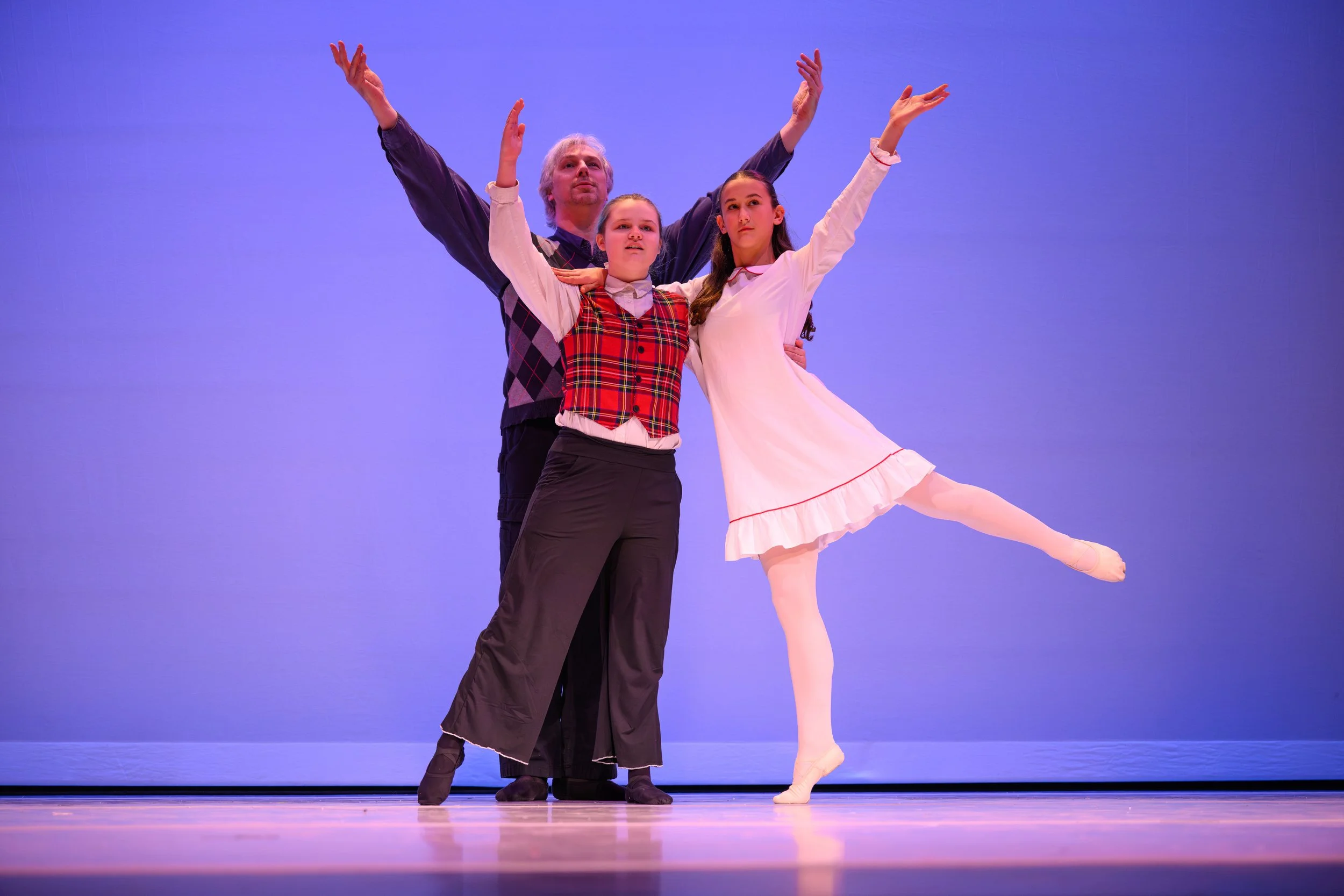 Three ballet dancers on stage, posing in ballet positions with arms raised, against a purple backdrop.