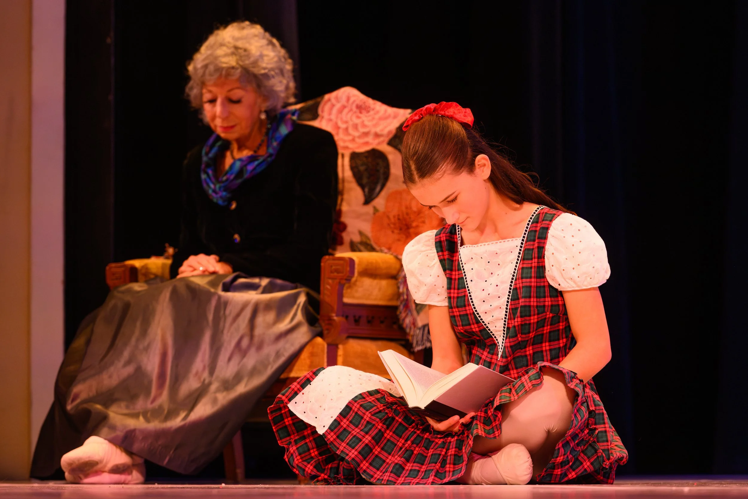 A young girl dressed in a plaid dress with puffy white sleeves, sitting on the floor reading a book, with an older woman sitting on a decorated armchair in the background.