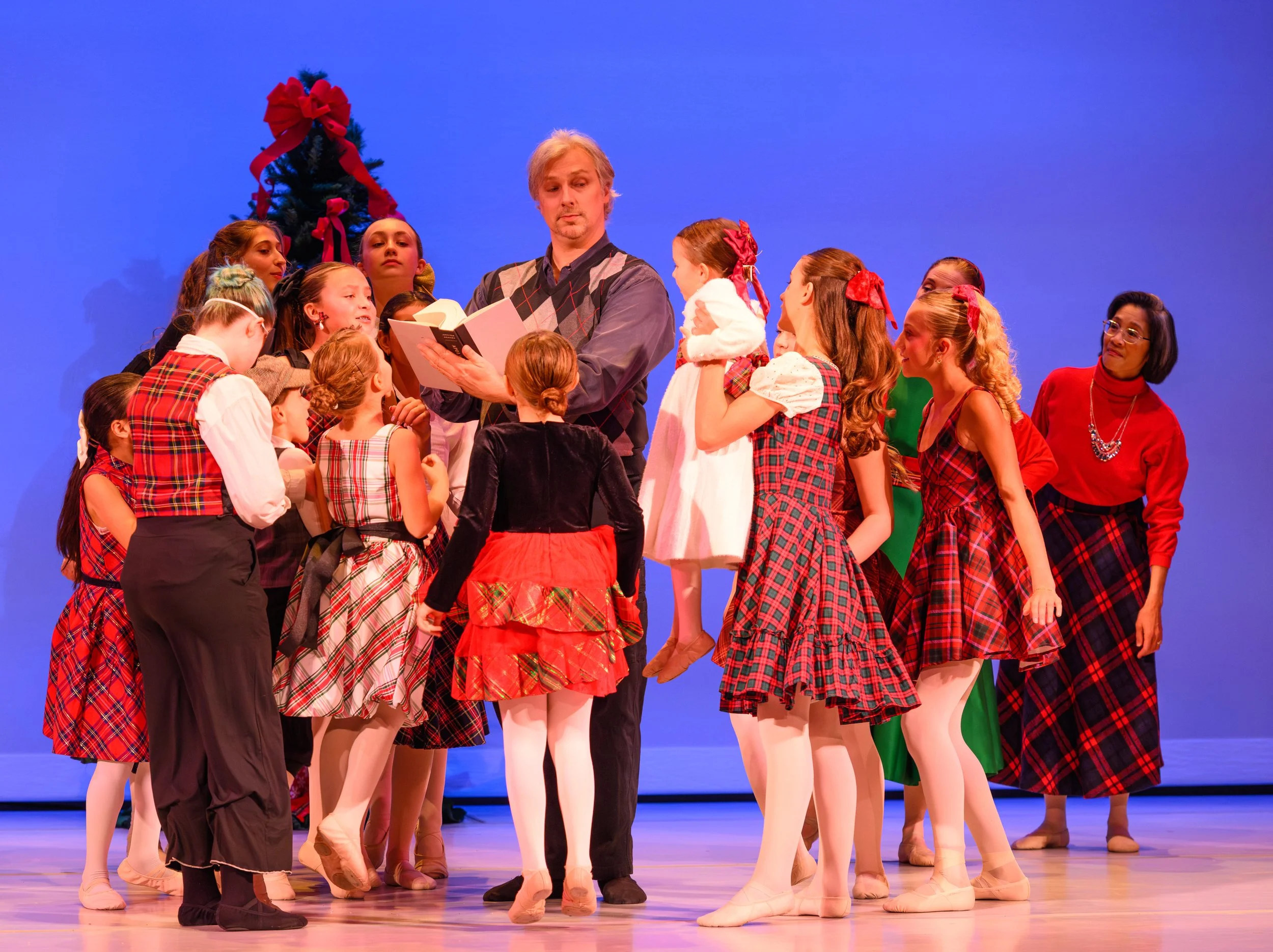 Children and an adult on stage during The Nutcracker Ballet, with a Christmas tree in the background, some children are wearing plaid costumes, and the adult holding a book.