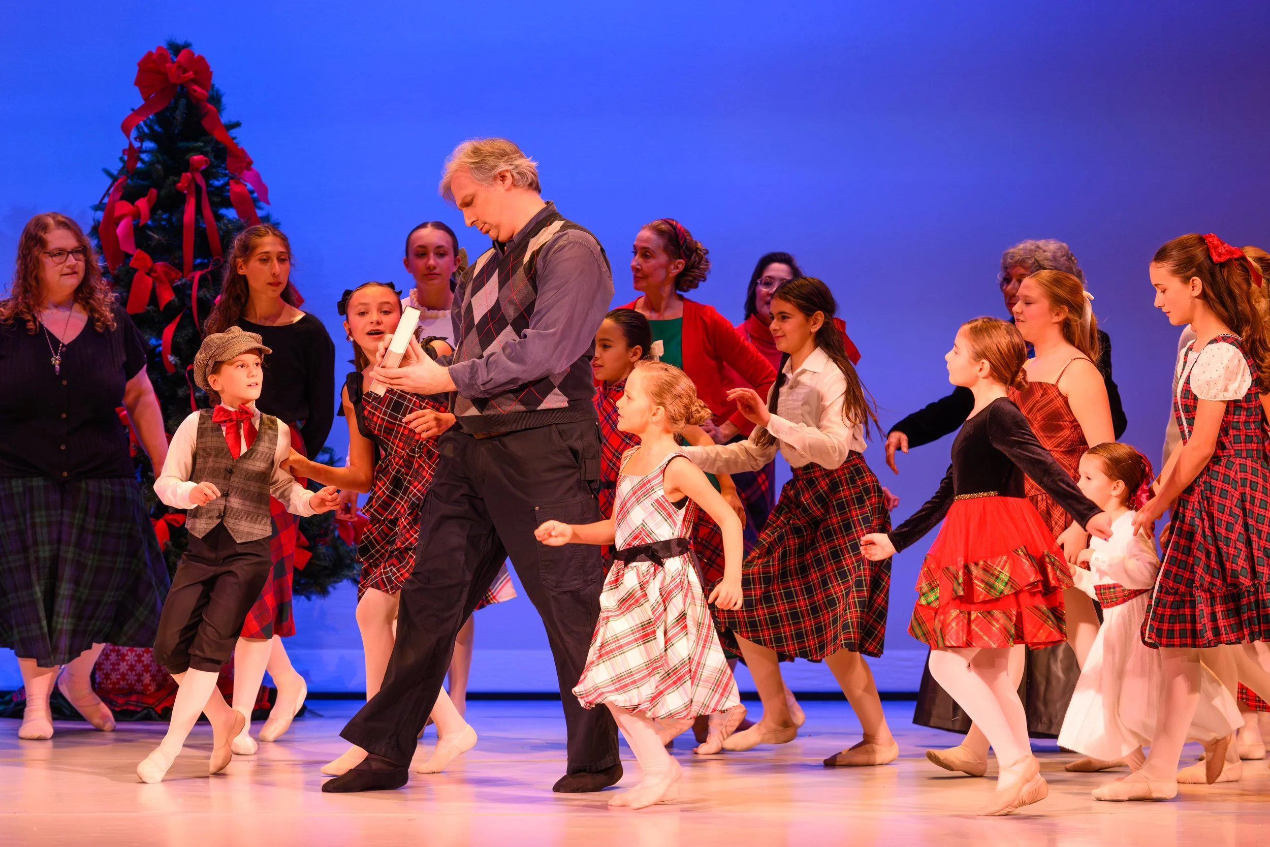 A group of children in tartan and Christmas-themed costumes dancing on stage with a man, in front of a decorated Christmas tree.
