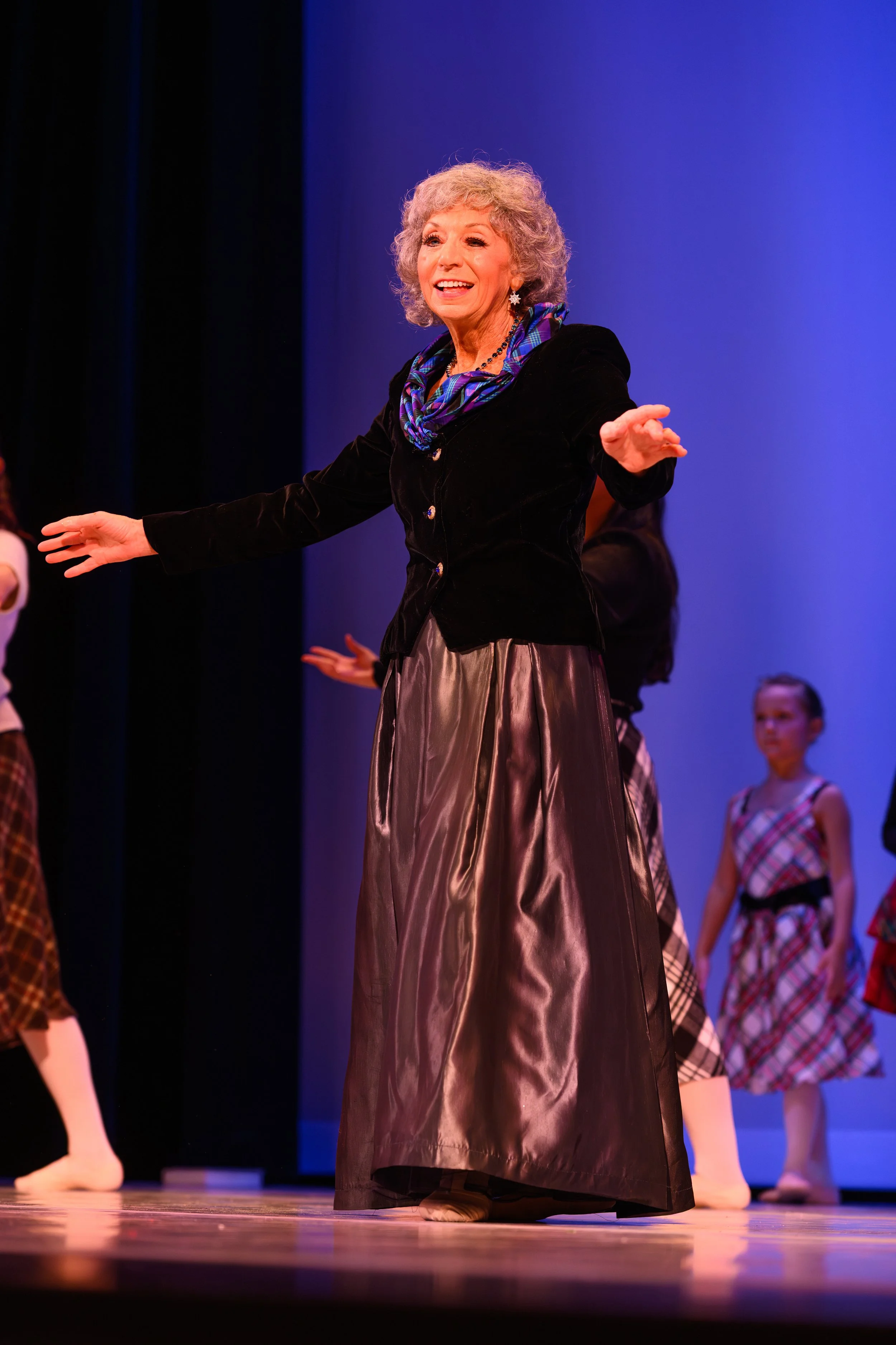 An elderly woman with curly gray hair, wearing a black velvet jacket, a blue plaid scarf, and a long satin skirt, performing on stage with children in plaid dresses in the background.