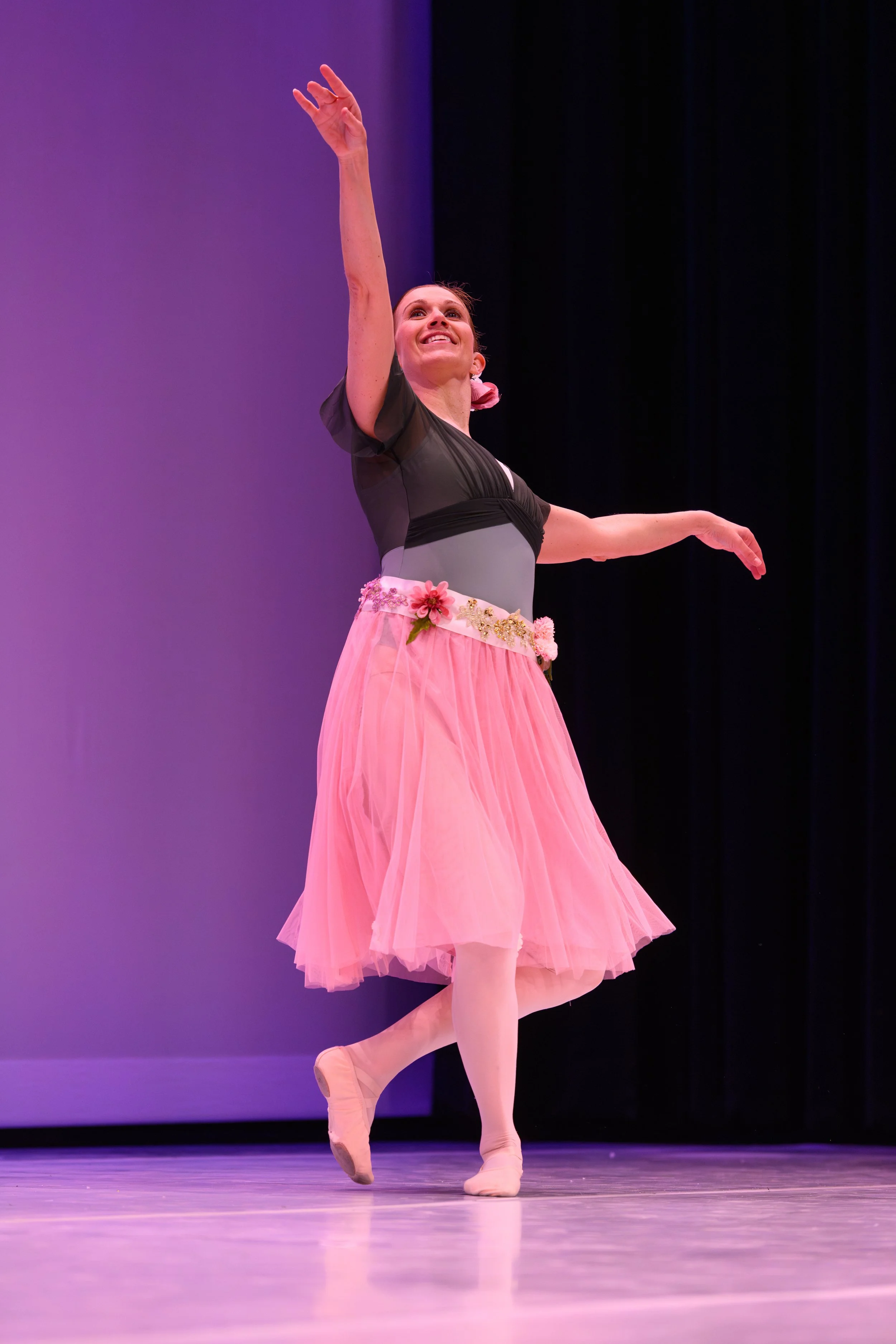 A ballerina performing on stage, wearing a pink tutu with floral decorations, a green and gray top, and pink tights, smiling and reaching upward with her right arm.