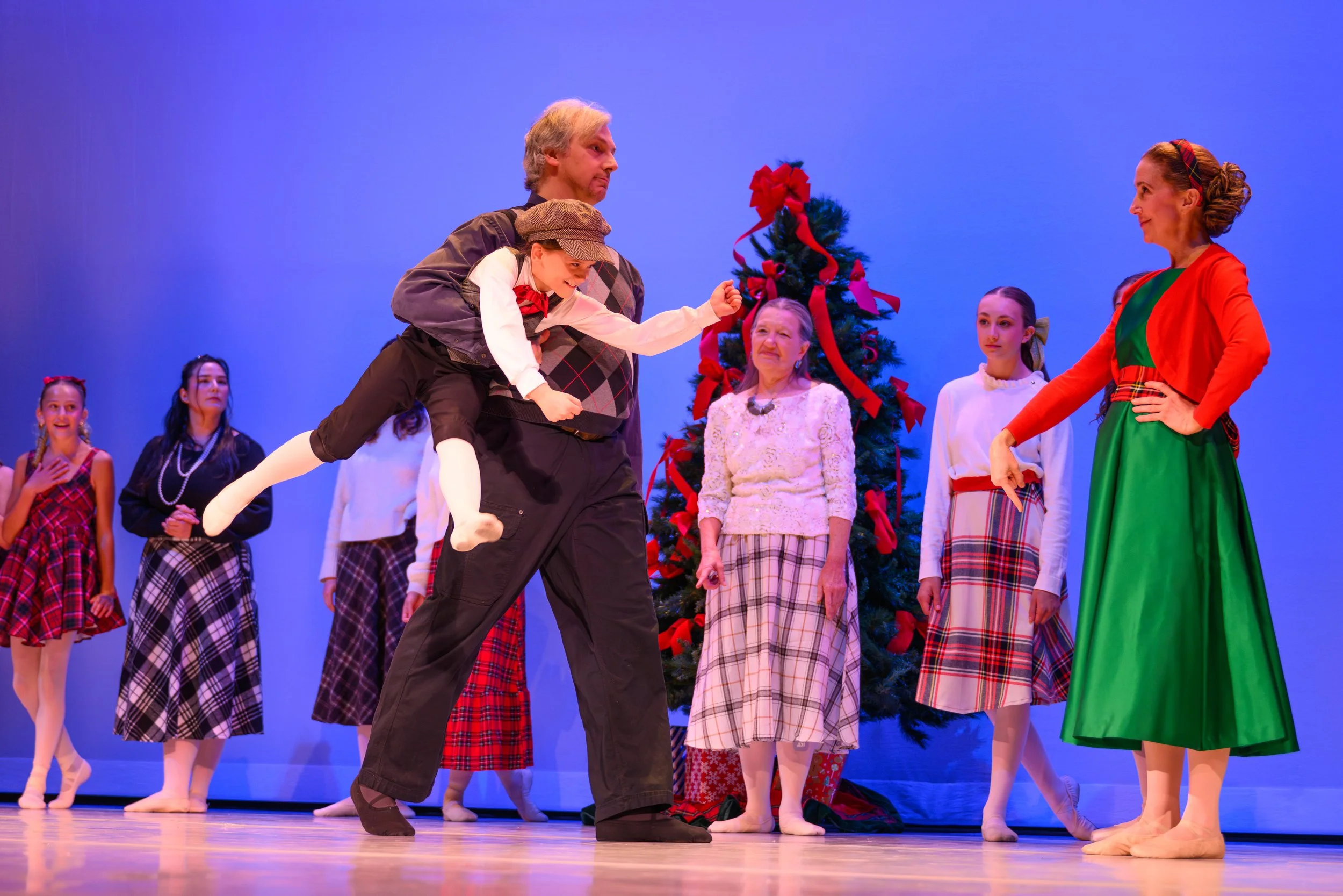 Children and adults dressed in festive costumes performing The Nutcracker Ballet with a decorated Christmas tree.