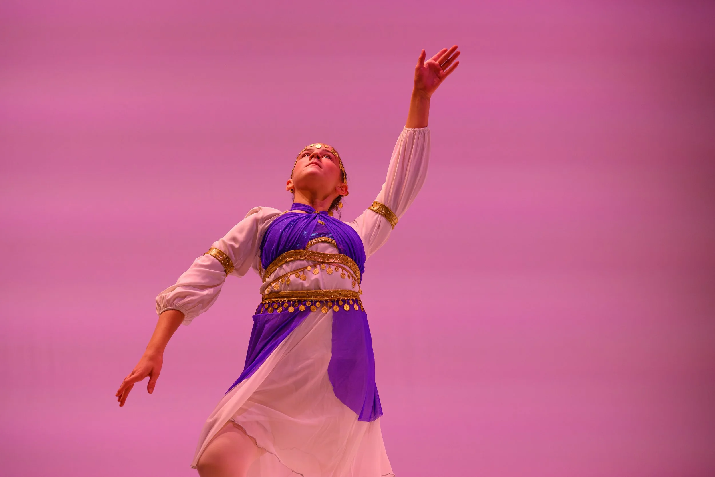 A young girl dressed in a flowing costume with gold embellishments and a purple sash, performing a dance with her right arm extended upward against a pink sky background.