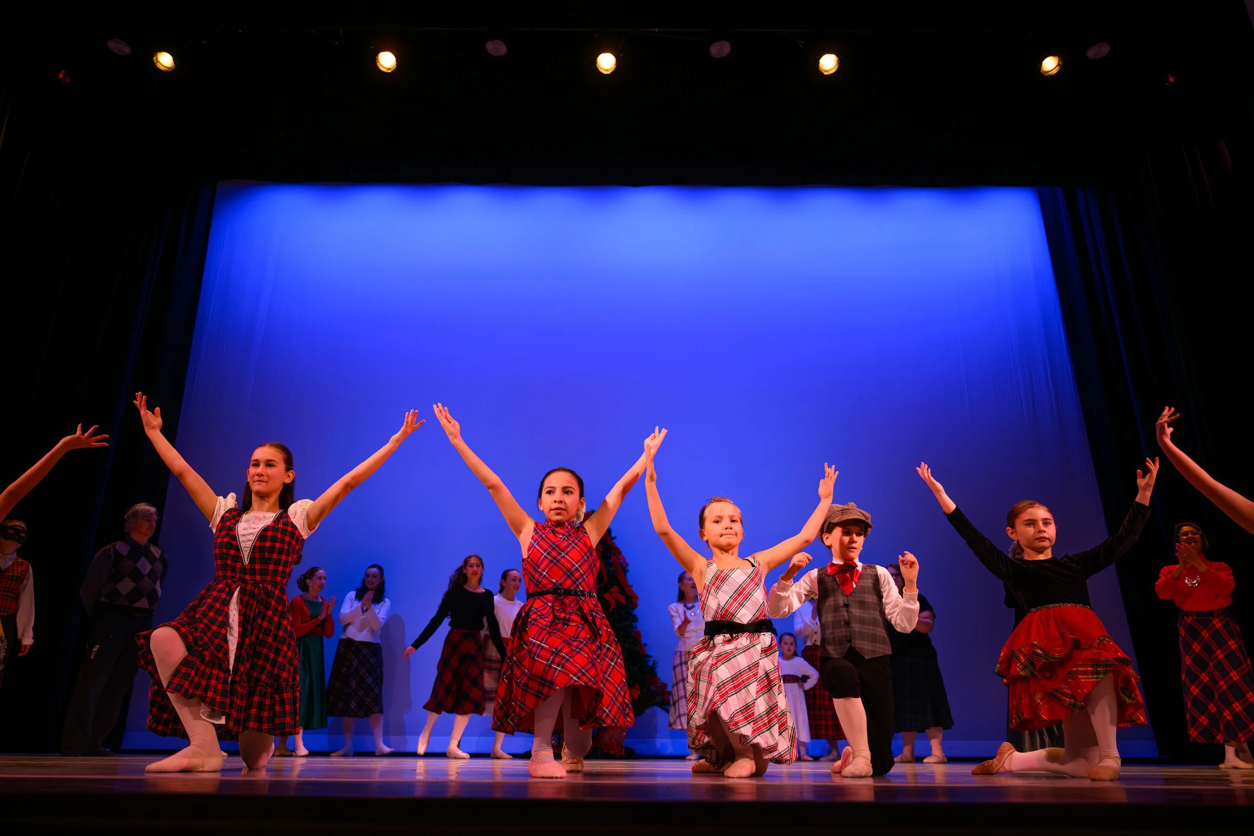 Children dressed in plaid and festive costumes performing a dance on stage with a blue backdrop.