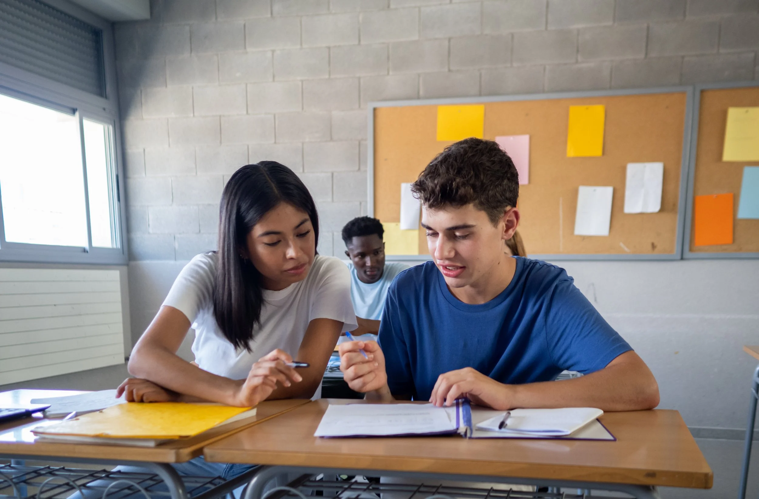 Group of three teenagers studying together in a classroom, sitting at a desk with notebooks and papers.