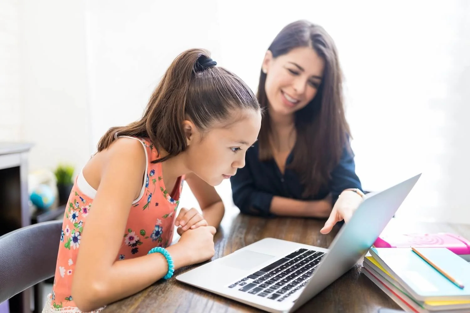 A woman and a young girl looking at a laptop together, sitting at a wooden table in a bright room with some books and school supplies on the side.