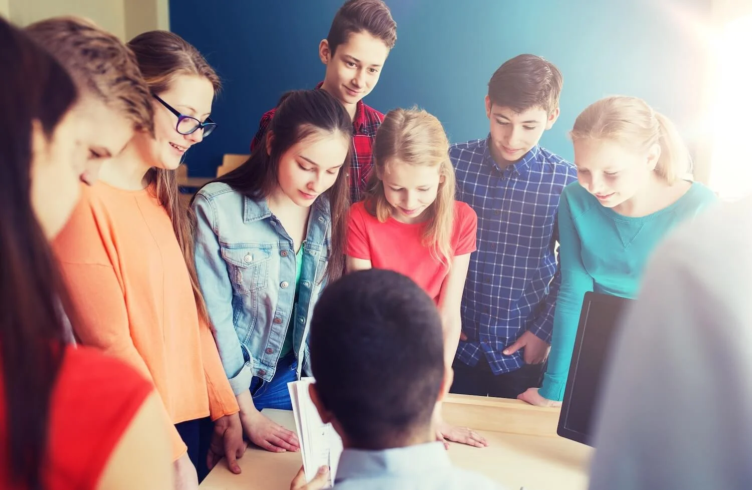 Group of seven young students gathered around a seated teacher in a classroom, looking at a book or tablet.