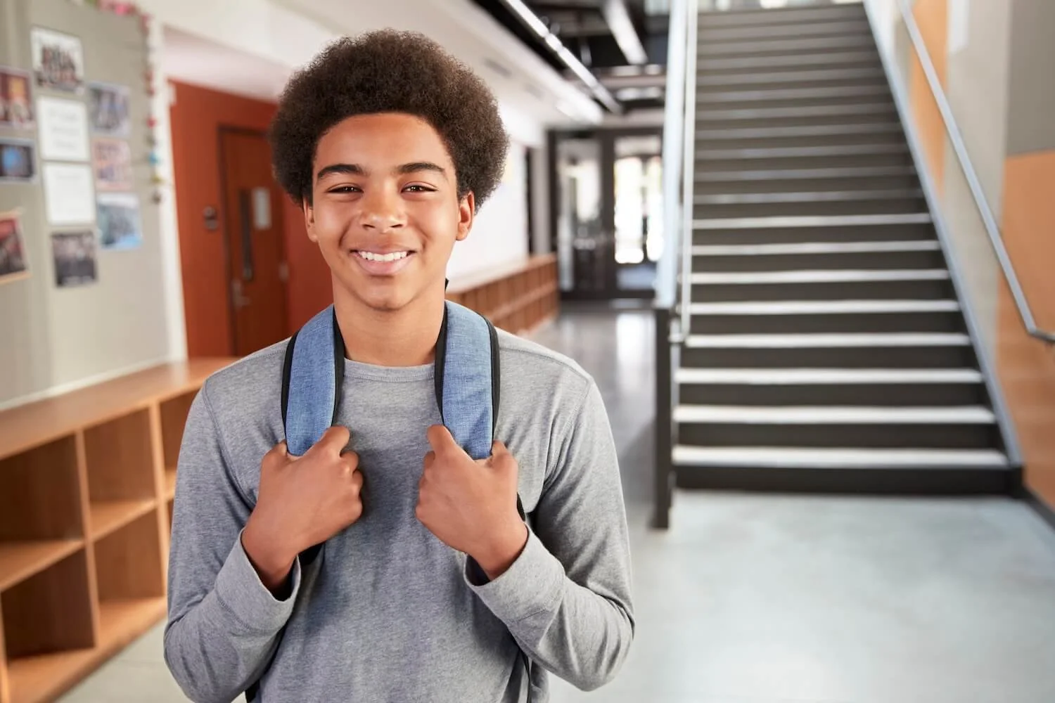 A smiling high school student standing in a hallway, holding the straps of his backpack