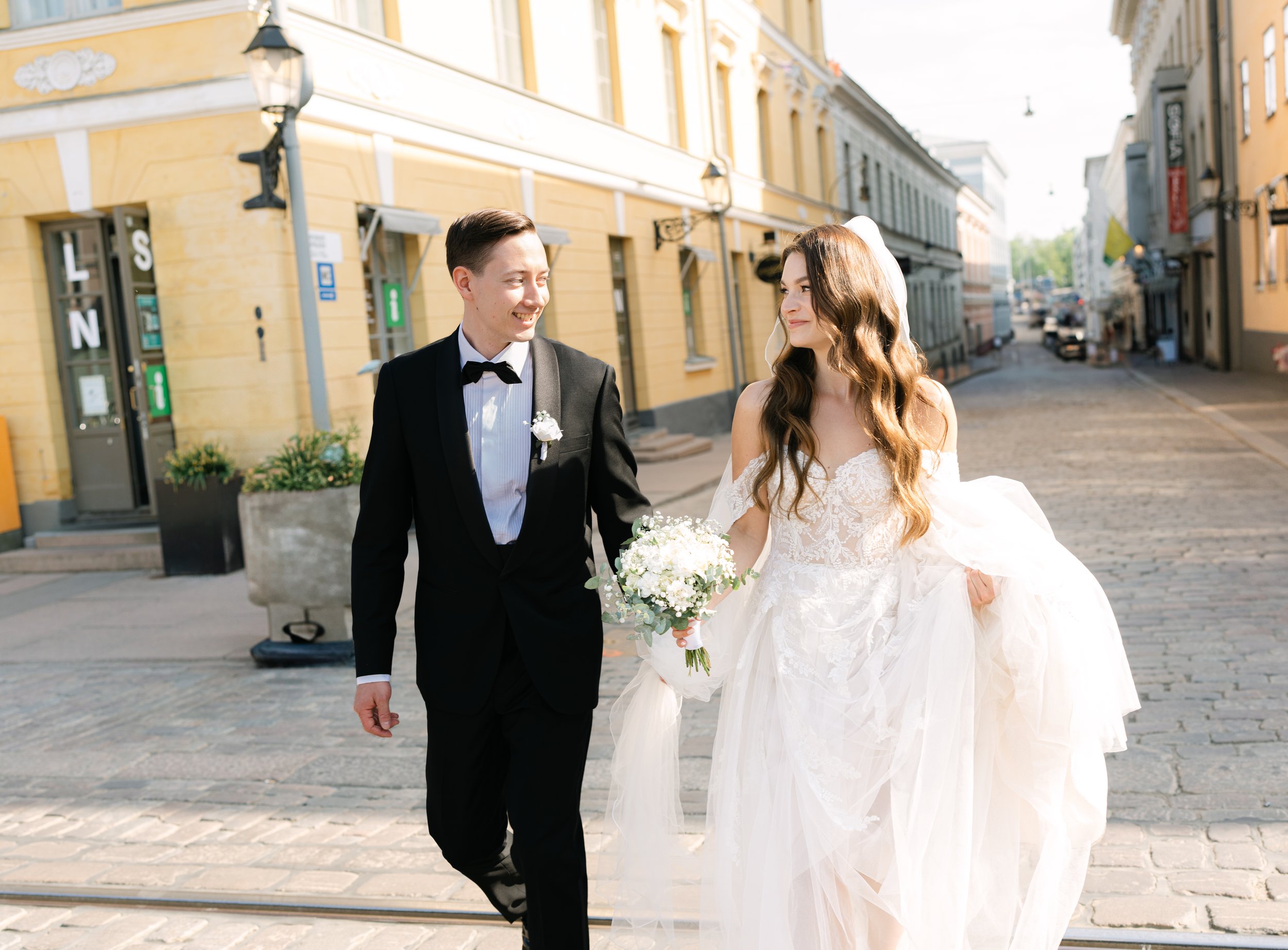 A bride and groom sitting on wooden stairs amid autumn foliage, embracing and sharing a tender moment, with the bride holding a bouquet of flowers.