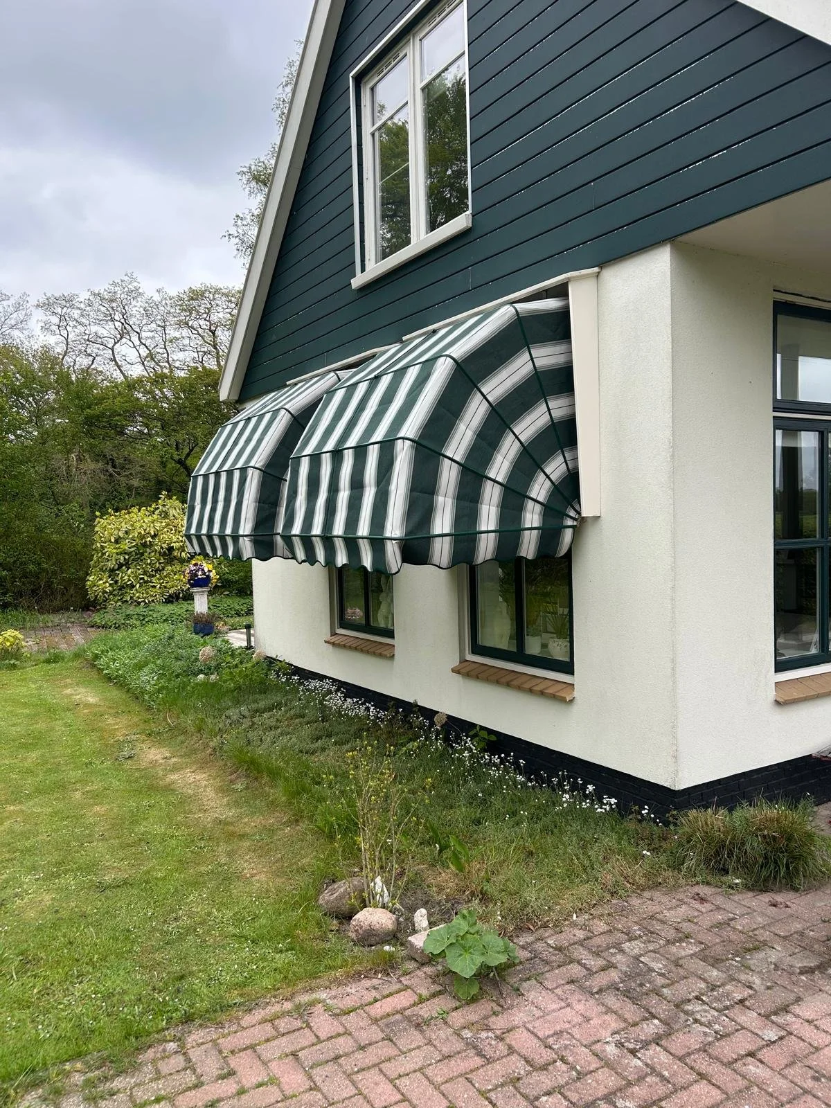 Een huis met een blauwe houten bovenverdieping en een witte onderverdieping, met uitzicht op een tuin. Er hangt een groene en witte gestreepte markies boven een raam aan de onderverdieping.