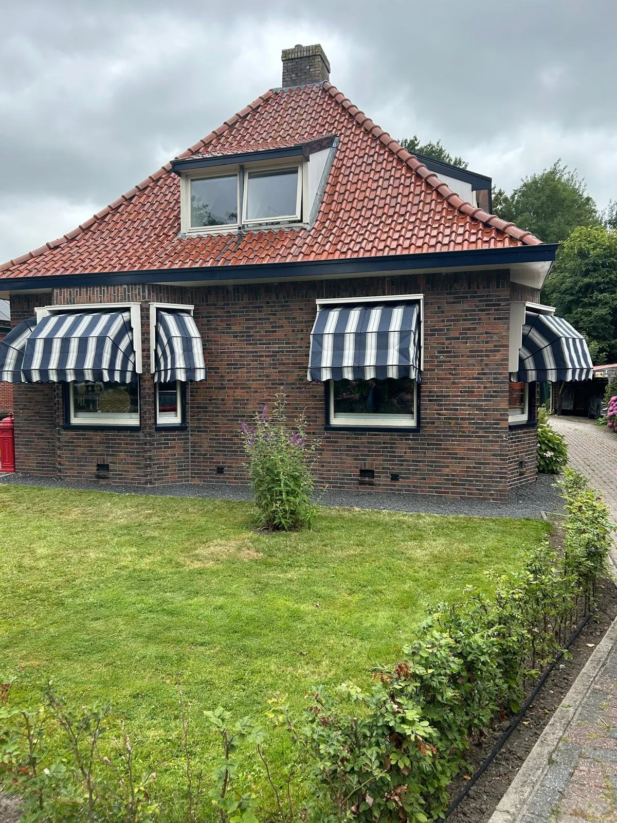 Huis met rode dakpannen, twee verdiepingen, en blauwe en witte gestreepte zonneluifels voor de ramen op de begane grond. Voor het huis ligt een gazon met een struik en een tuinpad links.