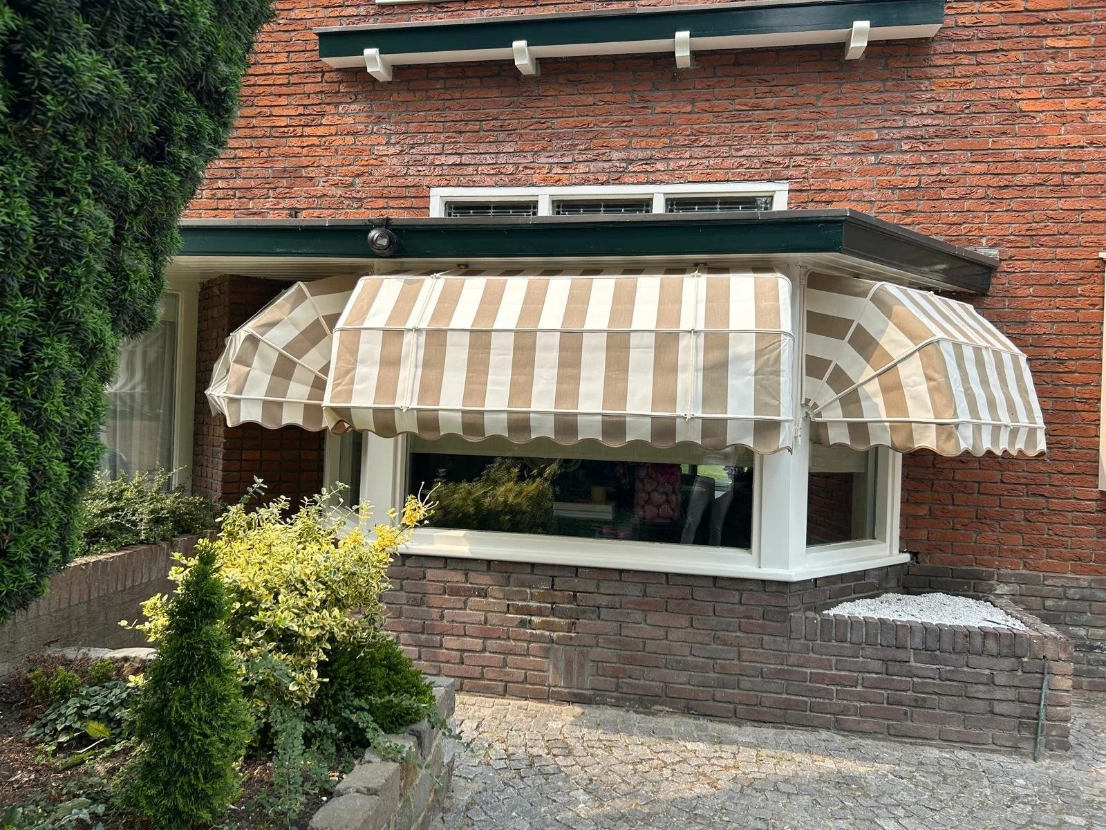 Buitenaanzicht van een huis met een grote ramen en een aftrekbare luifel met beige en witte strepen, rondom planten en een stenen oprit.