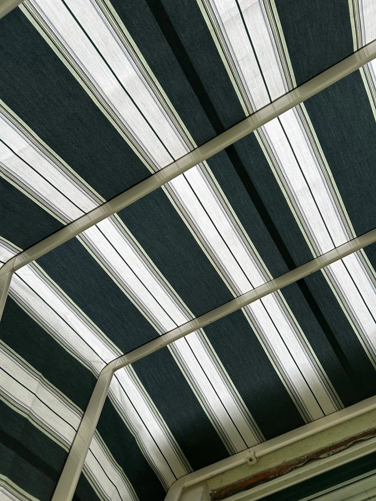 Het plafond van een overdekt terras met een stoffen luifel met zwarte, witte en grijze strepen, ondersteund door witte metalen balken.
