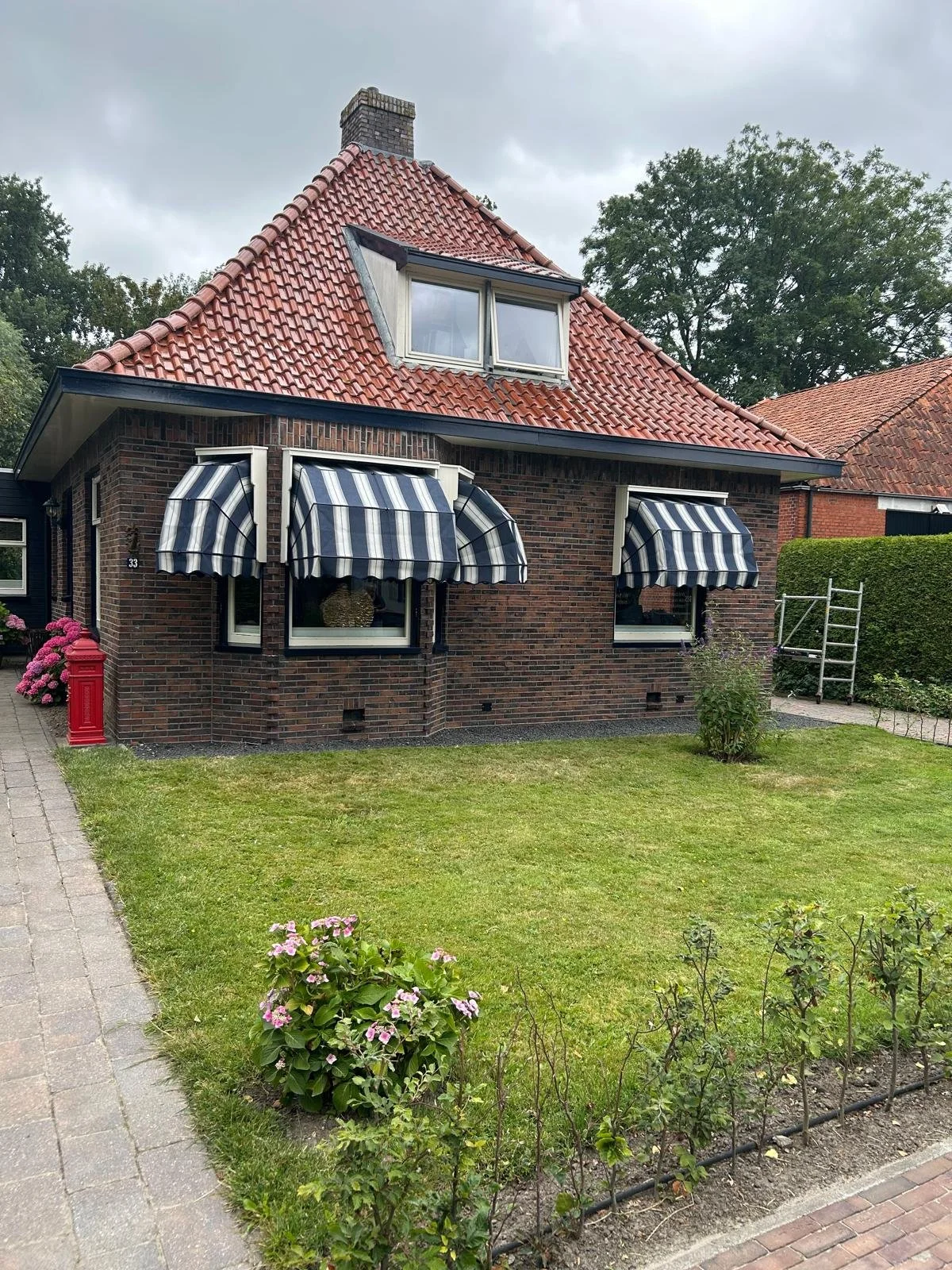 Een stenen huis met een rood tegeldak, blauwe en witte gestreepte uitbouwoverkappingen bij de ramen, een groene tuin en een ladder.