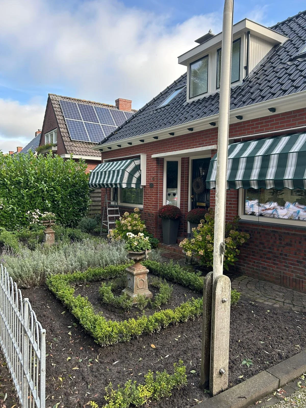 Een tuin met bloemen en struiken voor een rood bakstenen huis, met een dak met zonnepanelen. Er staan twee grote plantenbakken bij de deur, en een ladder staat tegen de muur. Een lantaarnpaal staat in het midden van het beeld. Er hangen markiezen.