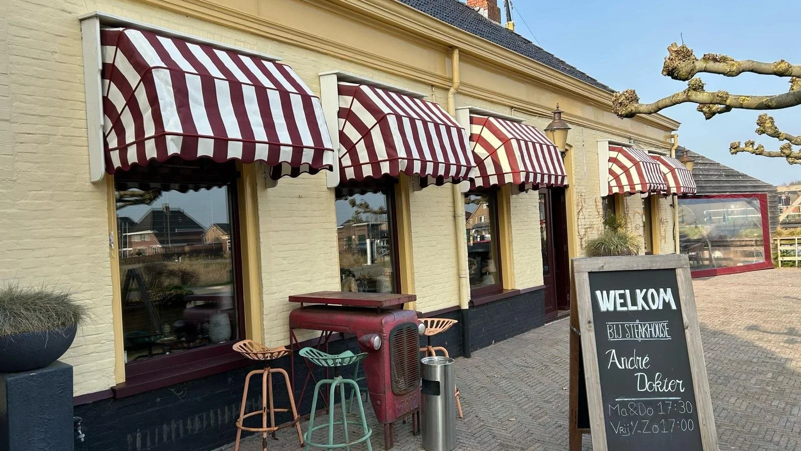 Een restaurant met rode en witte gestreepte markiezen, een zwart bord met witte tekst bij de ingang, buitenmeubilair, en een glazen windscherm op een terras.