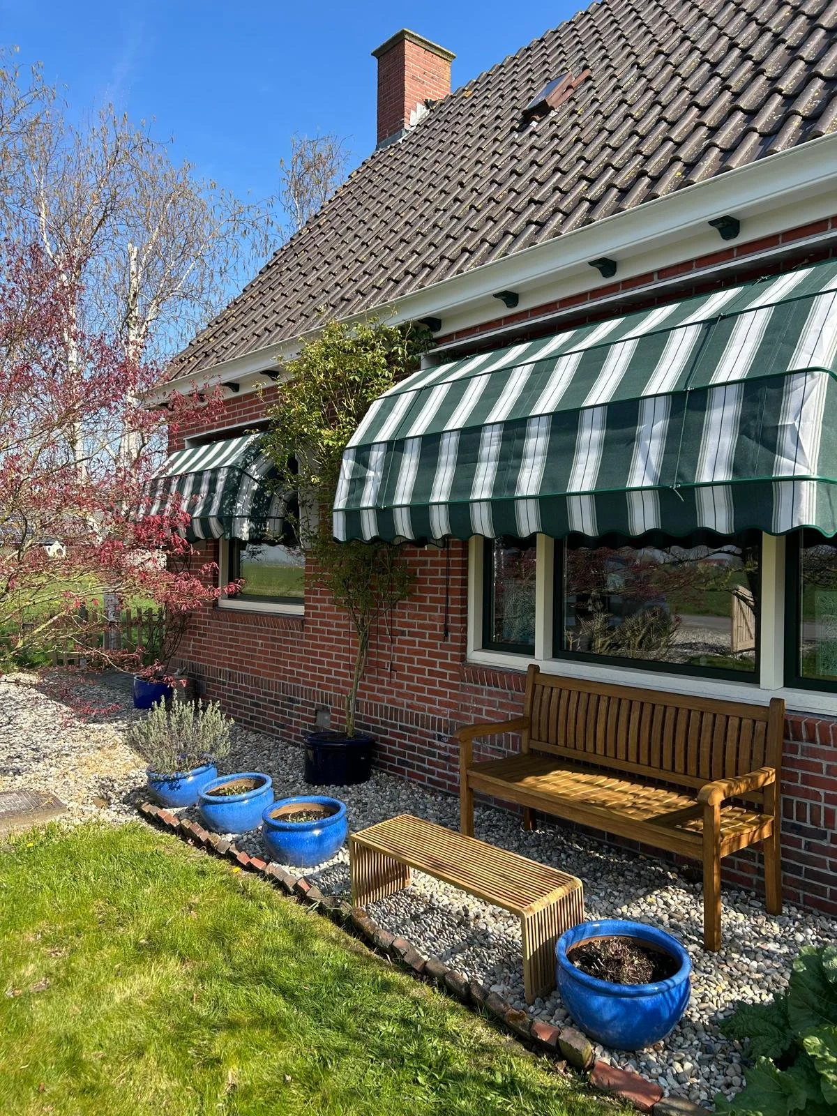 De gevel van een huis met ramen, groene en witte gestreepte markiezen, een bank en paar potten met planten in de tuin.