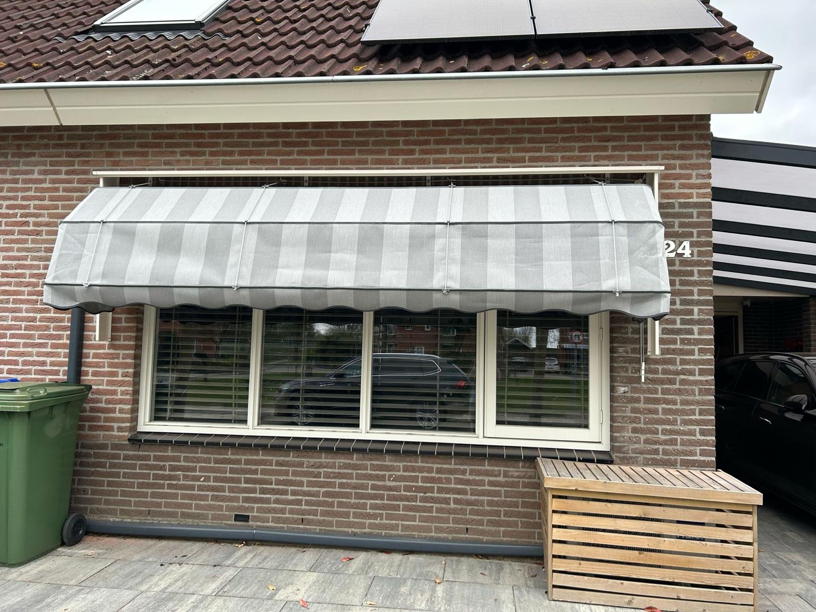 Bovenaanzicht van een huis met een dak met zonnepanelen en een uitgevouwen markies voor een groot raam, daarnaast een groene vuilnisbak en een houten bank.