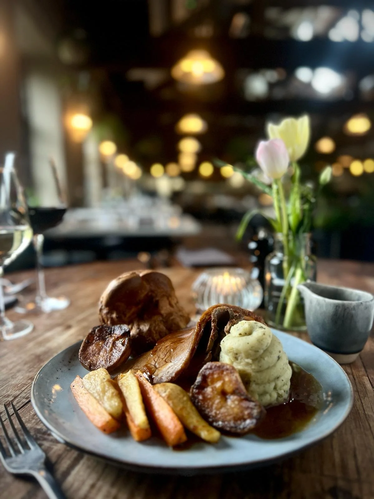 Plate of roasted meat with mashed potatoes, fried vegetables, and mushrooms on a rustic wooden table in a cozy restaurant setting with soft lighting and a flower arrangement in the background.
