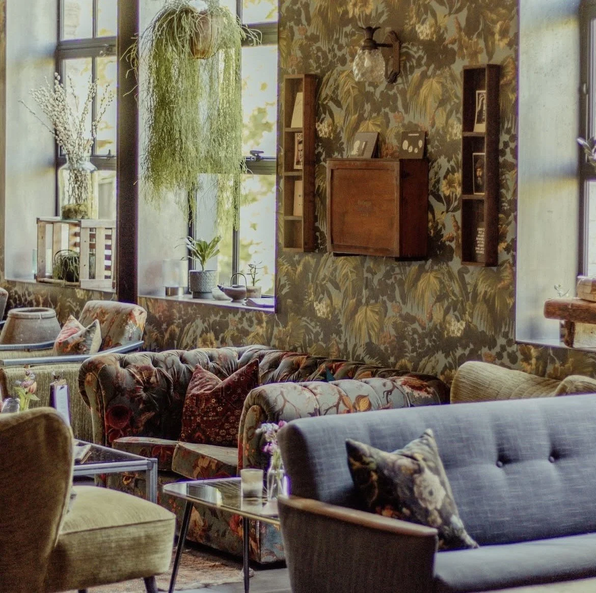 Cozy, artistic and vintage-style bar area with floral wallpaper, plush armchairs, a sofa, and shelves surrounding many antique tables with table candles and colourful flowers on top.